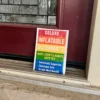 Deluxe Inflatable Roommate novelty sign with rainbow design and humorous features list displayed on a front doorstep.