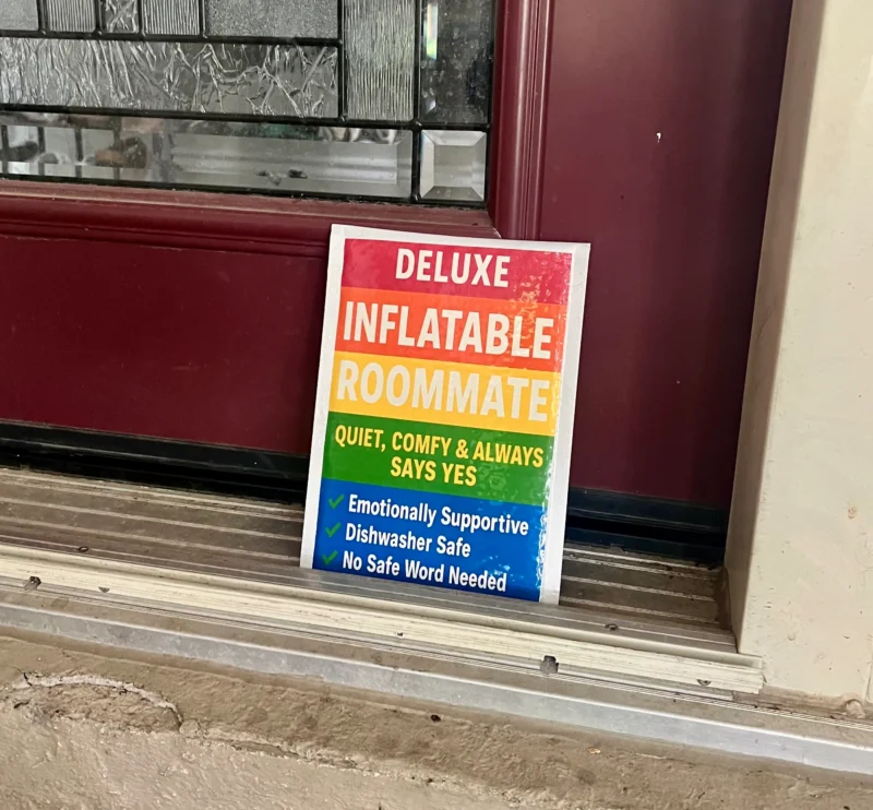 Deluxe Inflatable Roommate novelty sign with rainbow design and humorous features list displayed on a front doorstep.