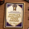 Framed “World’s Smallest Penis Award” prank certificate in gold and purple design displayed on a wooden desk.