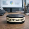 A hockey puck with the text "YOUR MESSAGE HERE" sitting on a desk in an office setting with a computer and phone in the background.