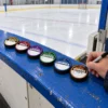 All six pucks lined up beside the bench Coach writing on a hockey puck award sticker beside five other award pucks on the boards at an indoor ice rink. Hardest Worker, Game Changer, My First Goal, Playmaker, Defensive Standout and Brick Wall stickers