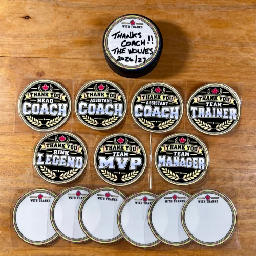Black and gold thank-you stickers for hockey coaches and team helpers, with white write-on backs on a wooden table. A puck above shows “Thanks Coach!! The Wolves 2026/27” handwritten on its sticker.
